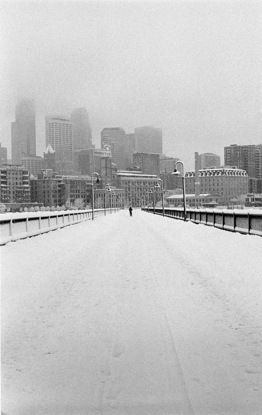 A person walks on a snow-covered bridge towards a city skyline shrouded in fog.