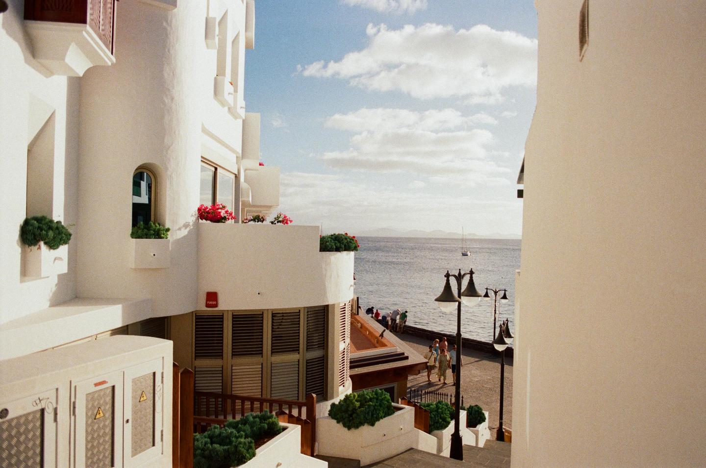 A white building with balconies and red flowers overlooks a body of water with a sailboat and distant mountains under a cloudy sky.
