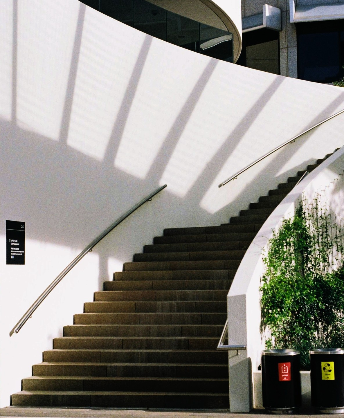 A curved outdoor staircase with a white wall and green foliage is illuminated by sunlight creating shadows.