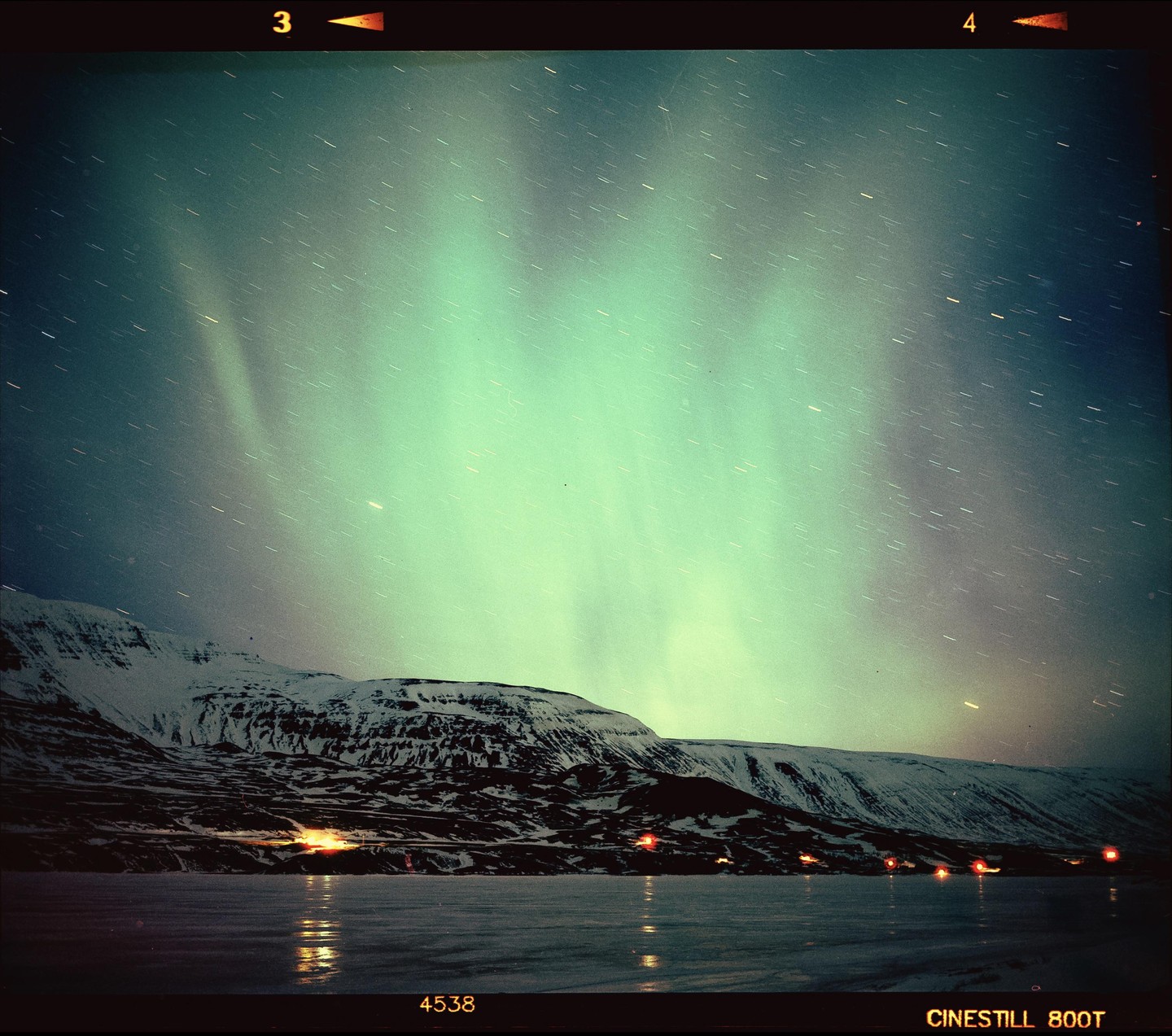 The aurora borealis glows green above a snow-covered mountain and a frozen lake with lights reflecting on its surface.