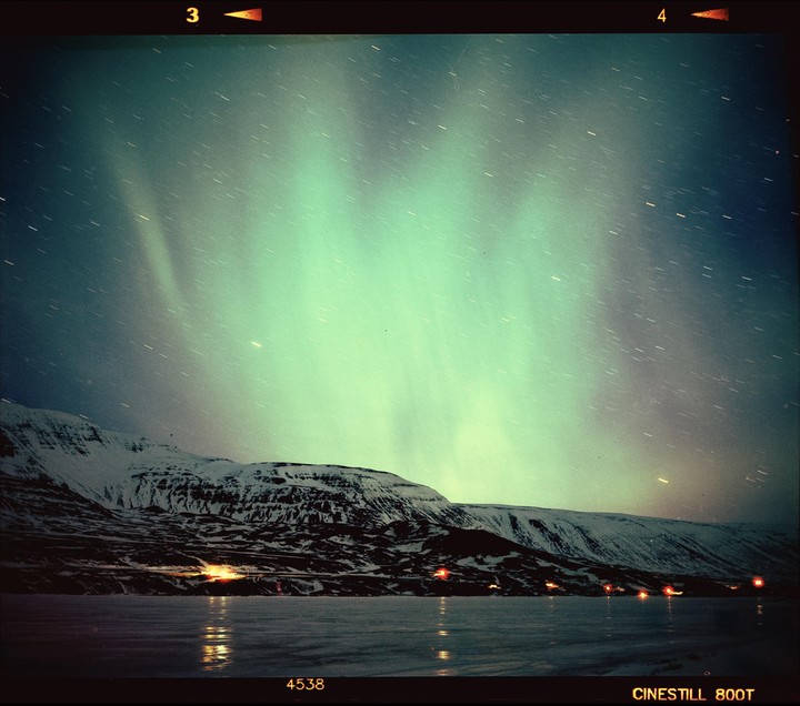 The aurora borealis glows green above a snow-covered mountain and a frozen lake with lights reflecting on its surface.
