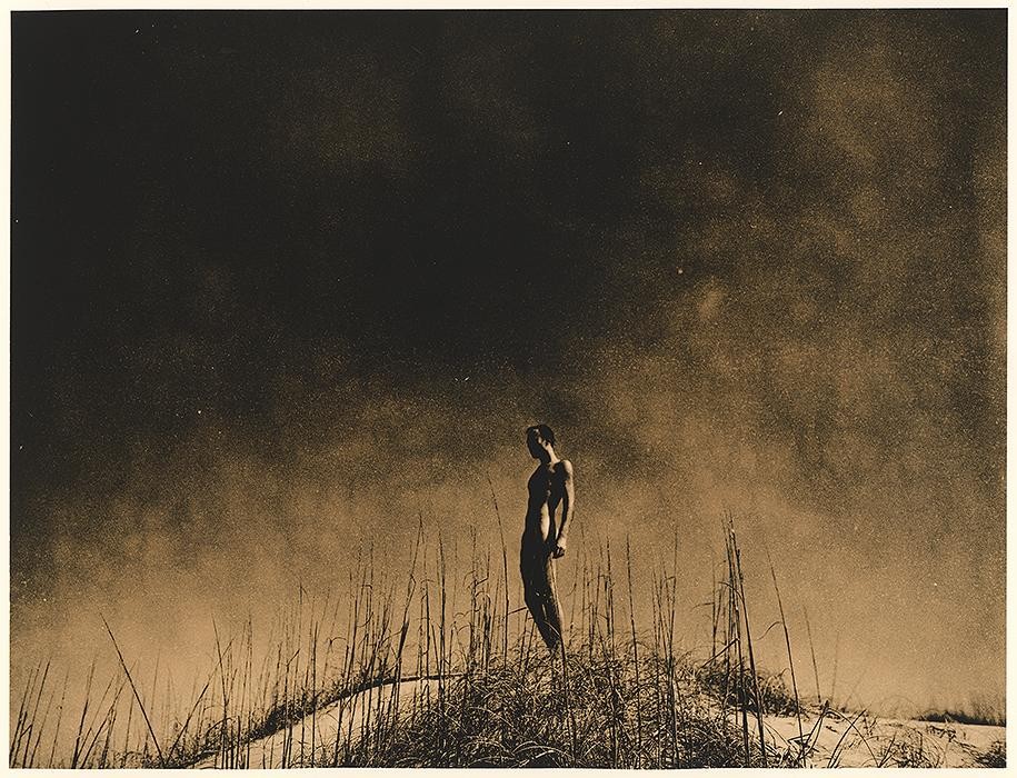 A nude man stands on a sand dune covered in tall grass under a dark, hazy sky.