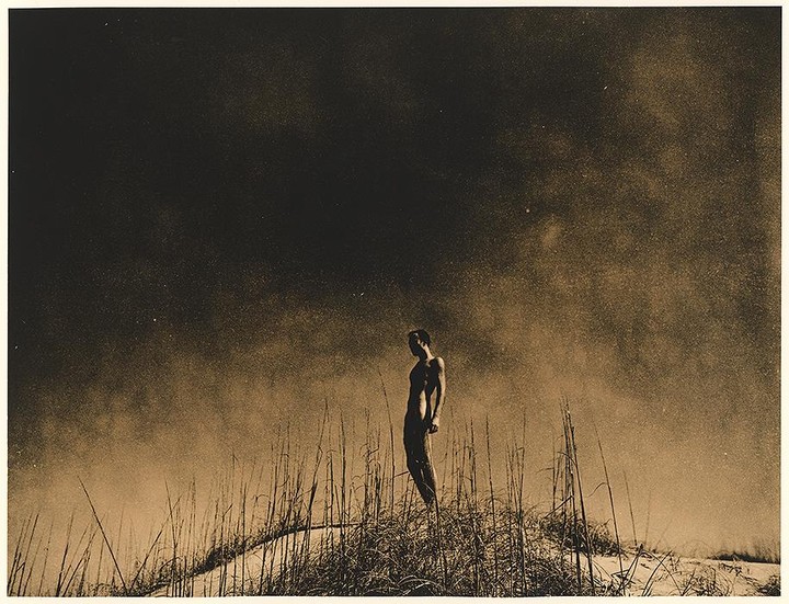 A nude man stands on a sand dune covered in tall grass under a dark, hazy sky.