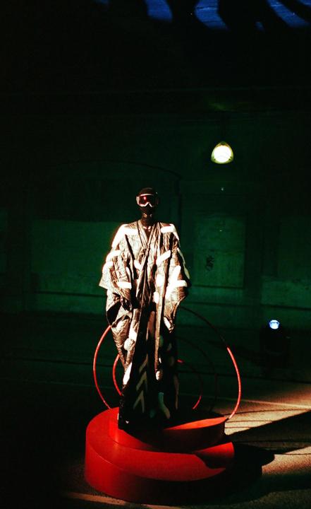 A mannequin wearing a patterned robe and goggles stands on a red platform in a dark room.
