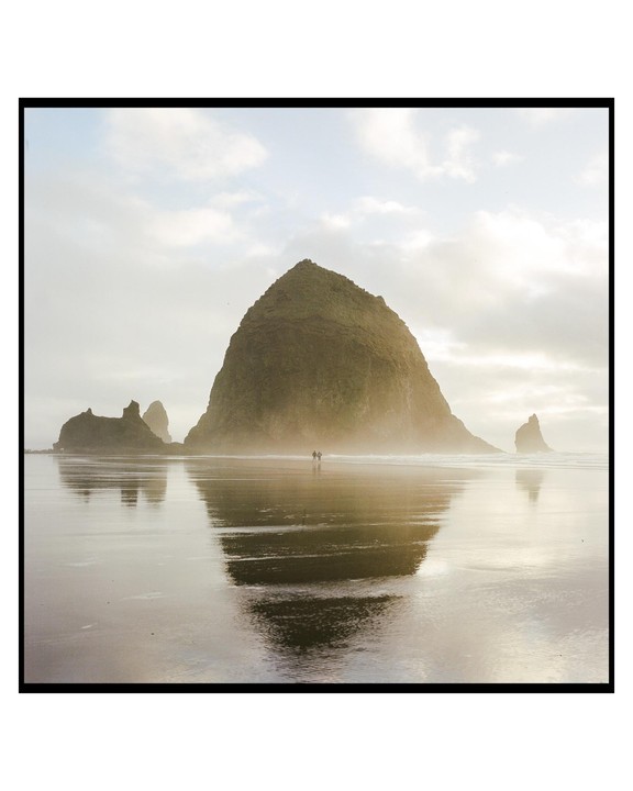 Two people walk on a wet beach with a large rock formation in the background, reflected in the water.