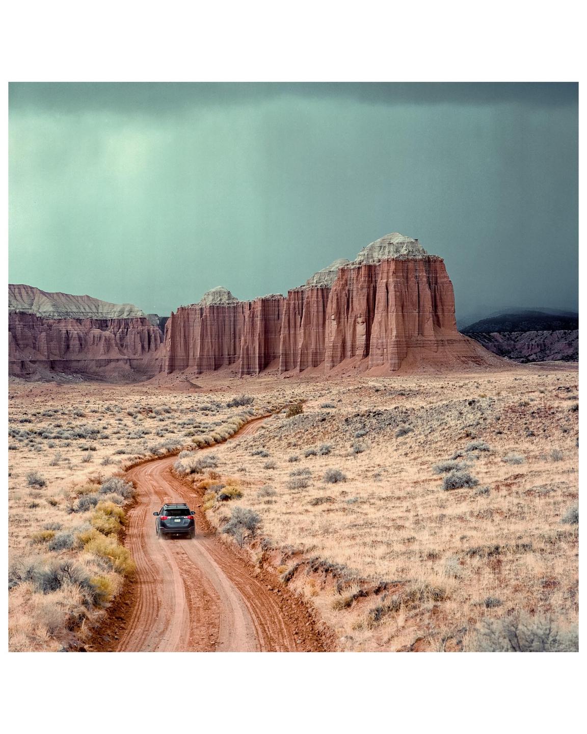 A car drives on a winding dirt road through a desert landscape with large rock formations under a cloudy sky.