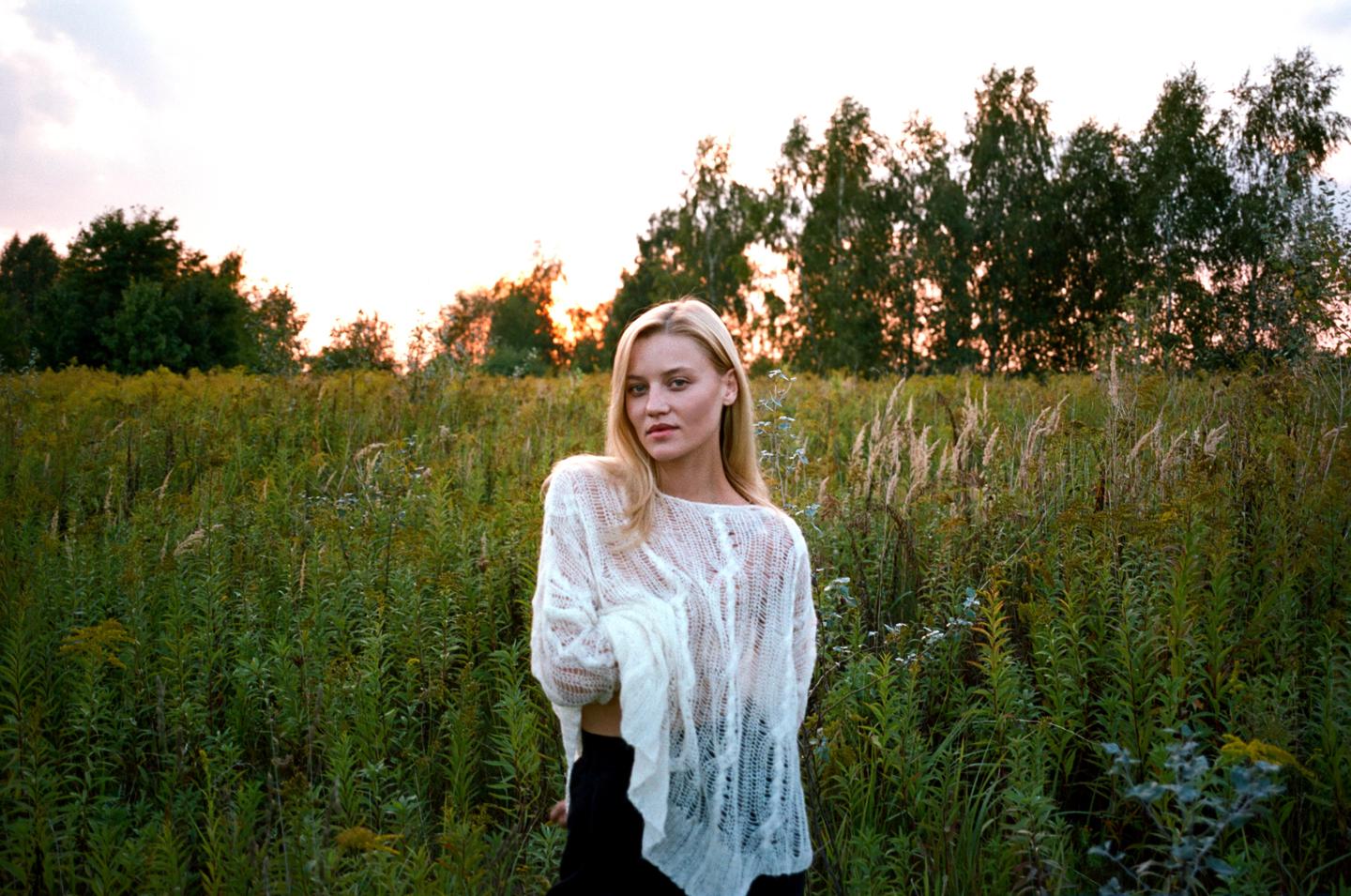 A woman stands in a field of tall grass and wildflowers with trees in the background at sunset.