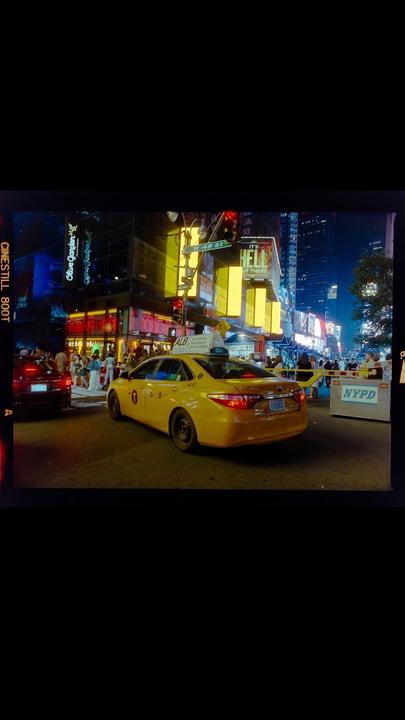 A yellow taxi cab drives down a busy street at night, surrounded by bright neon signs and tall buildings.