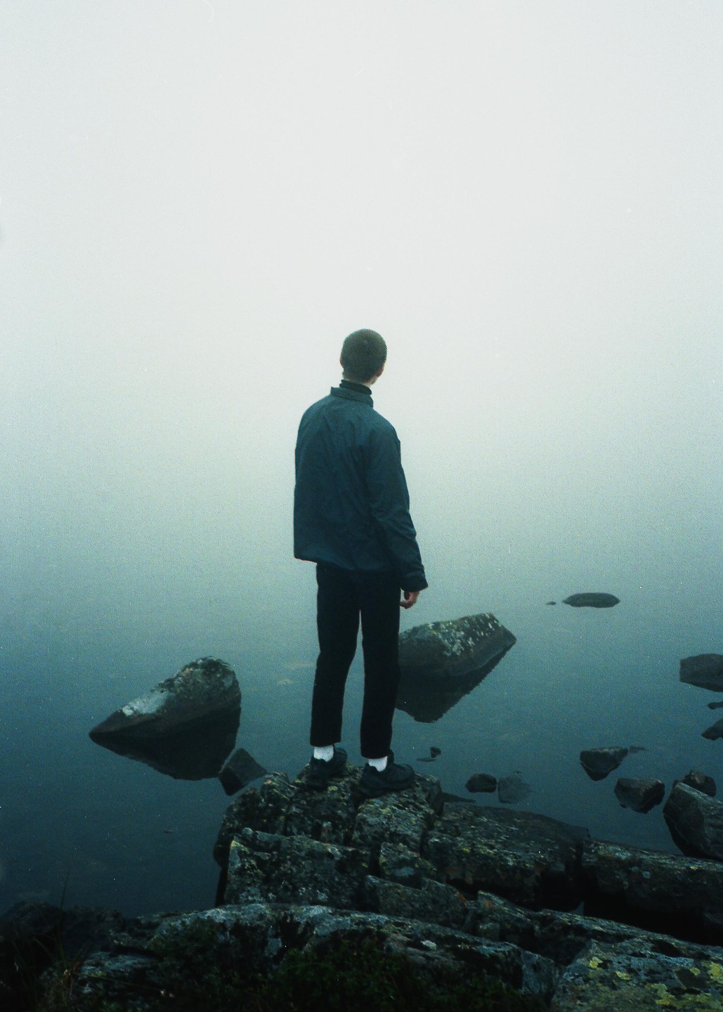 A man stands on a rocky shore looking out into a foggy body of water.