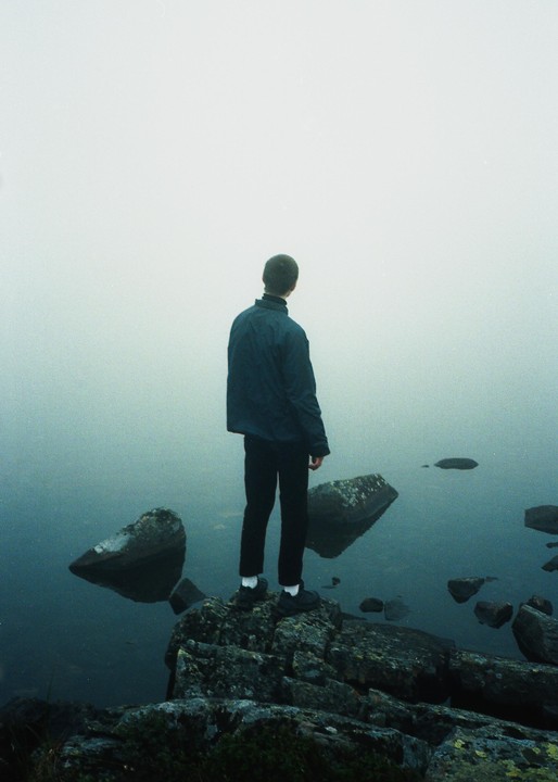 A man stands on a rocky shore looking out into a foggy body of water.