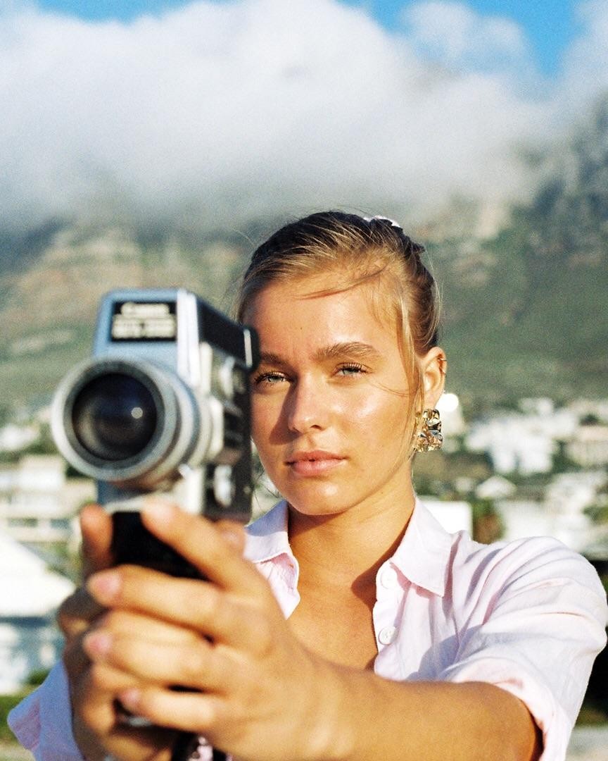 A woman holds a video camera toward the viewer with a mountain and cloudy sky in the background.