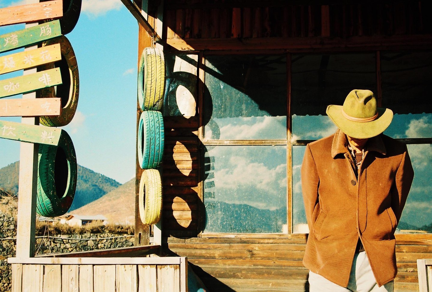 A man in a cowboy hat and brown coat stands in front of a wooden building with painted tires hanging on it.