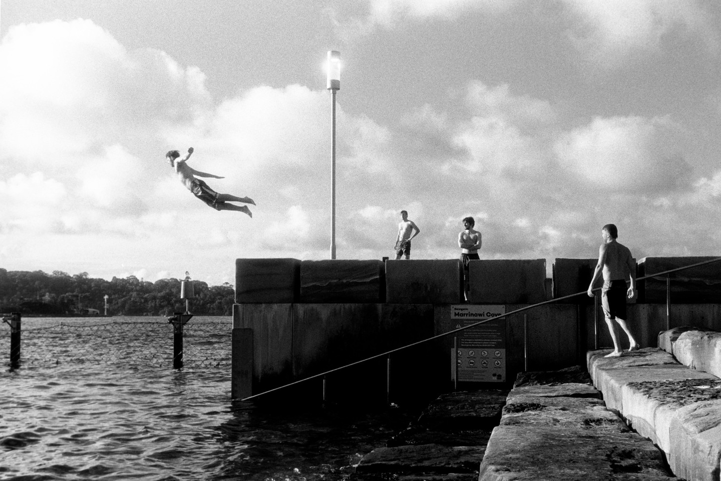 A man jumps into the water from a concrete wall while other men stand nearby under a cloudy sky.