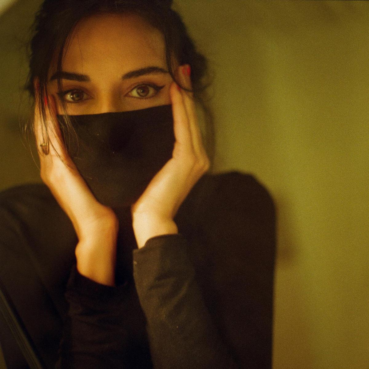 A woman with dark hair and red fingernails covers her face with her hands, revealing only her eyes.
