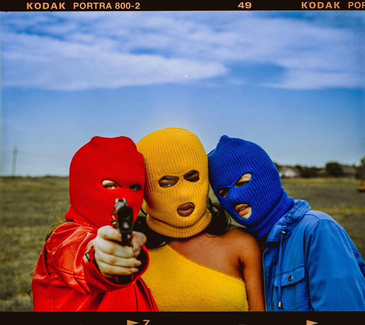 Three people wearing balaclavas pose for a portrait, one pointing a gun at the viewer.