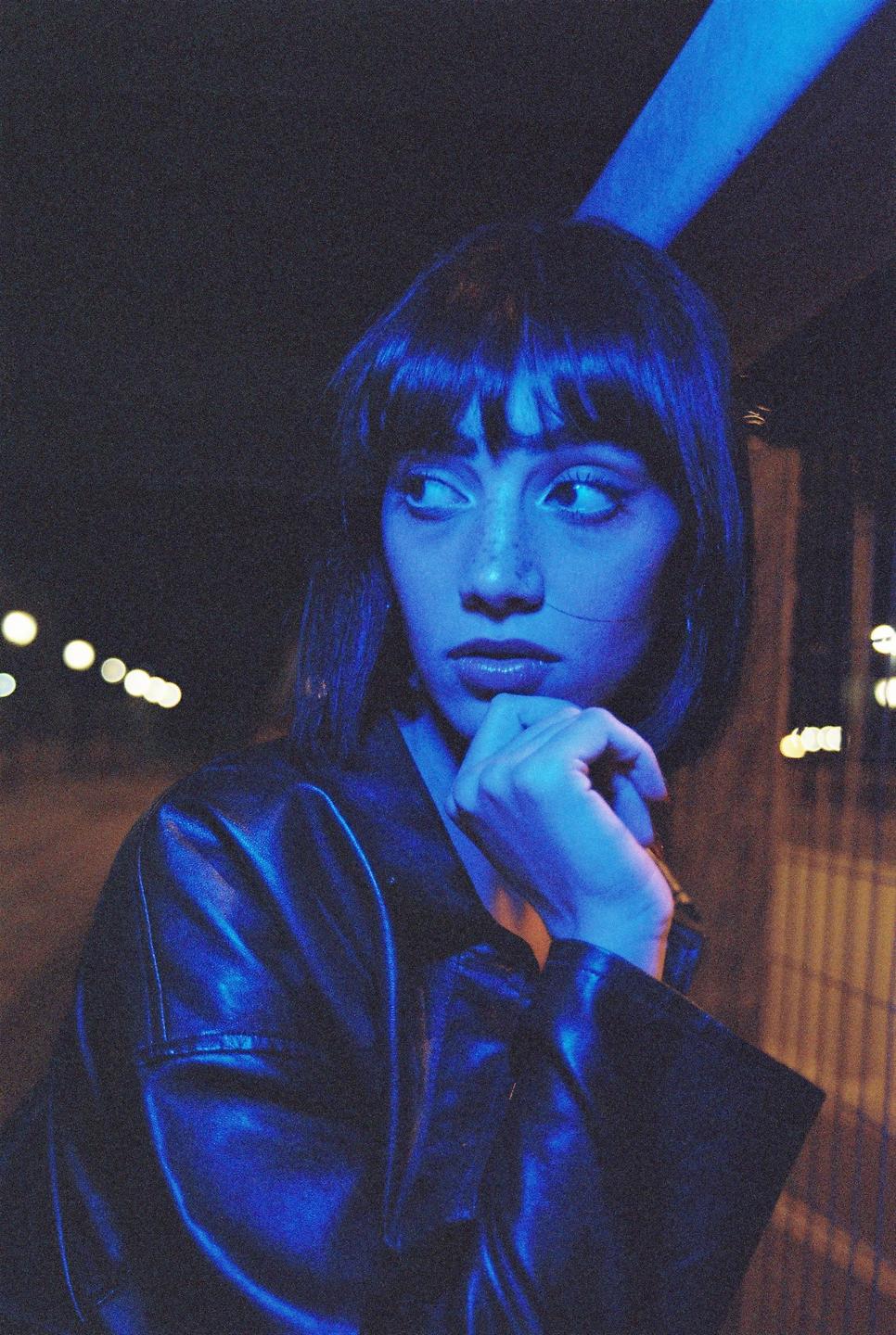 A woman with short hair and bangs looks to the side, illuminated by blue light at night.