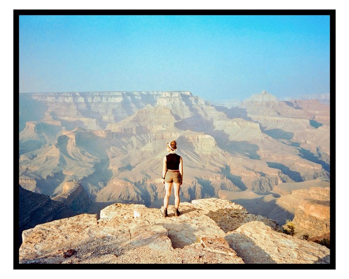 A woman stands on a cliff overlooking a vast canyon landscape under a clear sky.