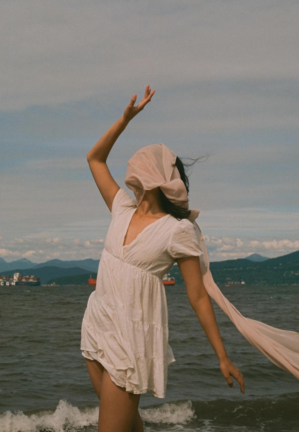 A woman in a white dress with a scarf covering her face stands by the ocean with mountains and ships in the background.