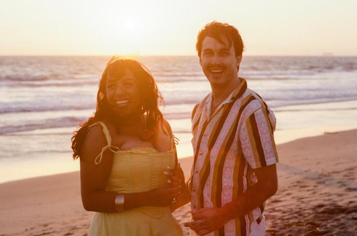 A couple stands on a beach at sunset, smiling at the camera.