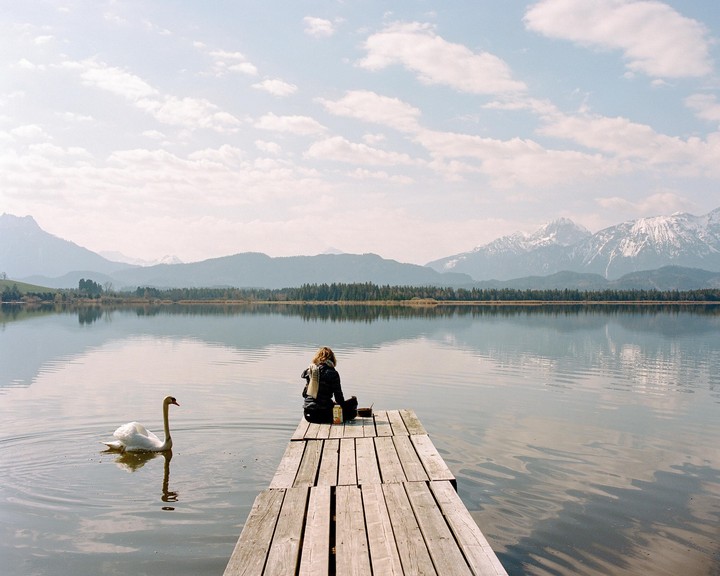 A woman sits on a wooden pier next to a swan on a lake with mountains in the background.