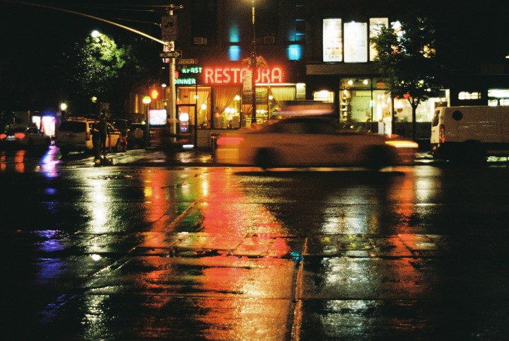 A blurred car drives past a restaurant with a neon sign on a wet street at night.
