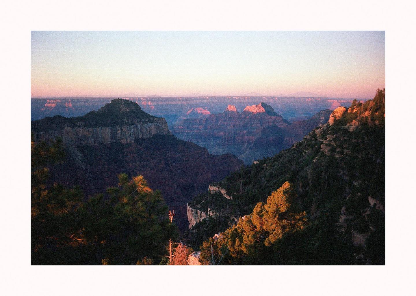 A vast canyon landscape is illuminated by the golden hour light, highlighting distant peaks and the rich colors of the rock formations.