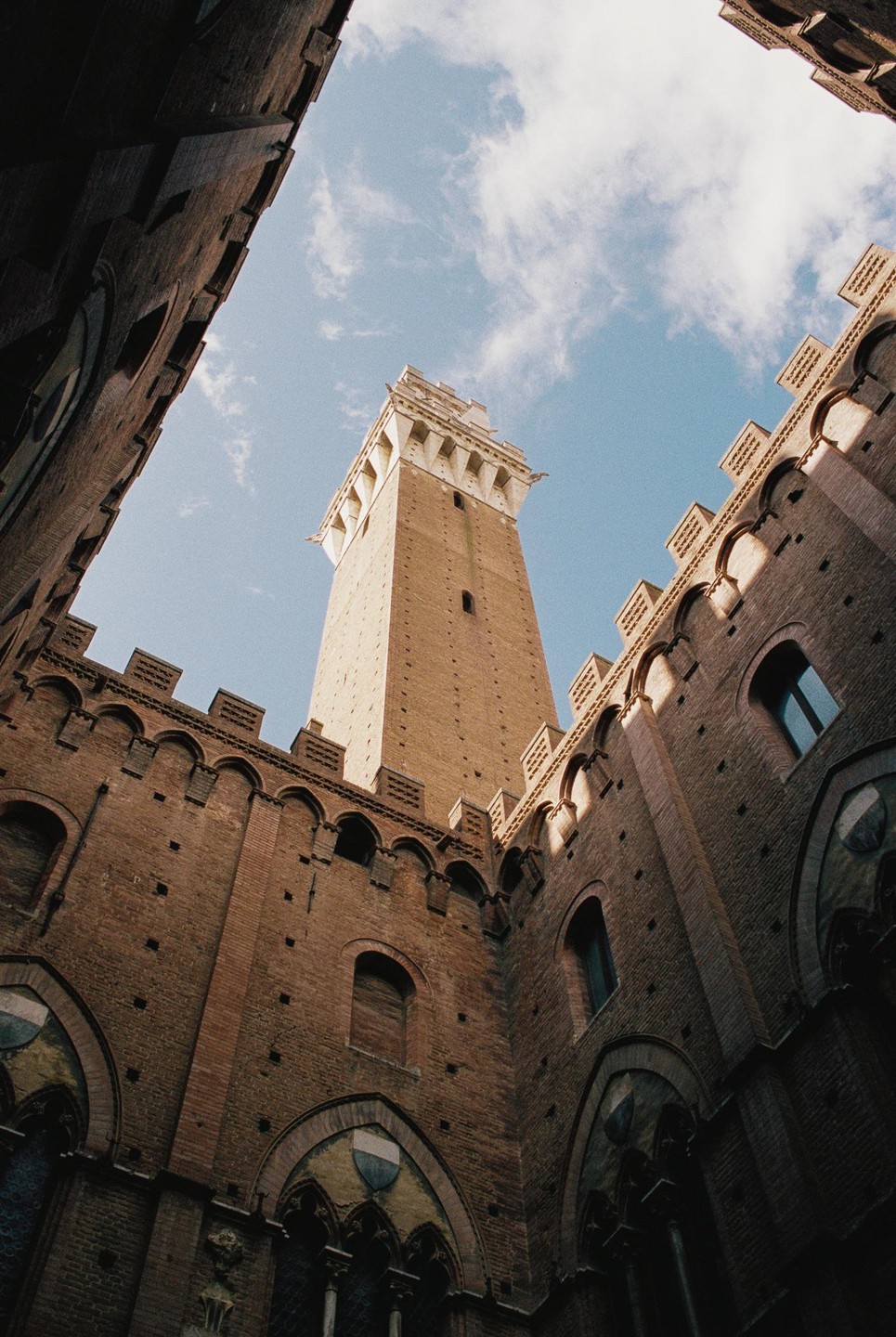 A tall brick tower rises above surrounding brick buildings with arched windows, all viewed from below against a blue sky with clouds.