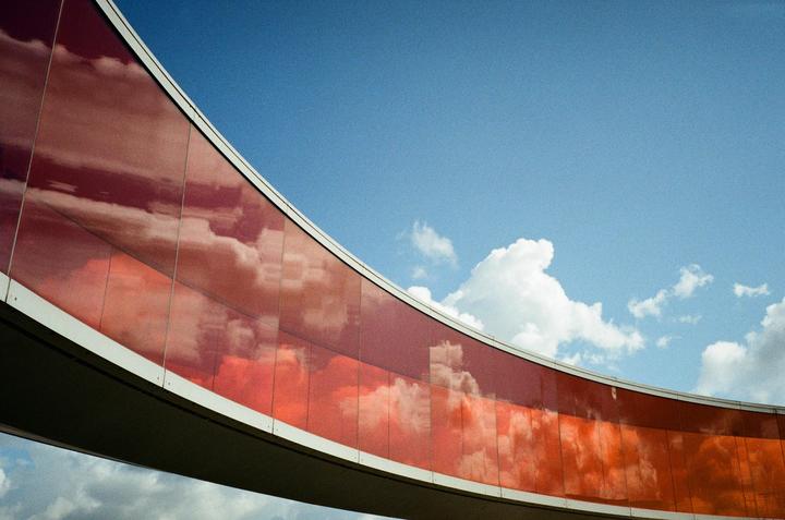 Clouds are reflected in the curved red glass of a modern building against a blue sky.