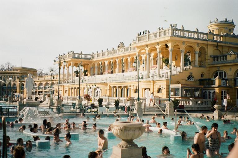 Many people are in a large outdoor swimming pool with a grand yellow building in the background.