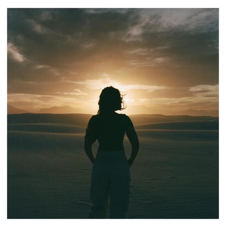 A woman stands silhouetted against a sunset over sand dunes and mountains.