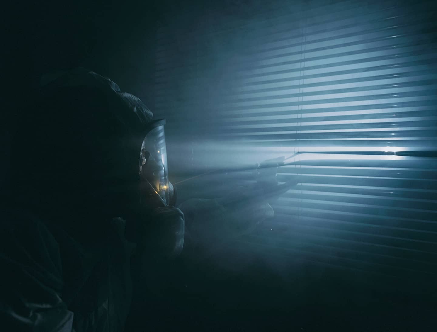 A person in a hazmat suit looks through window blinds into a bright light.