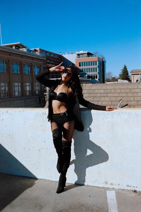 A woman in sunglasses, a black bra, and black shorts poses on a rooftop with buildings in the background.