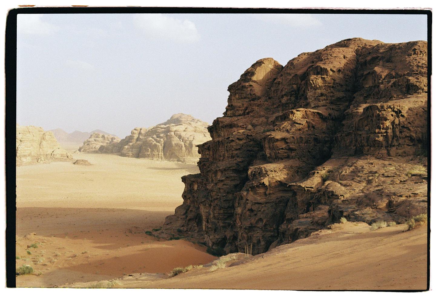 A vast desert landscape features a large rock formation on the right and smaller formations in the distance under a pale sky.