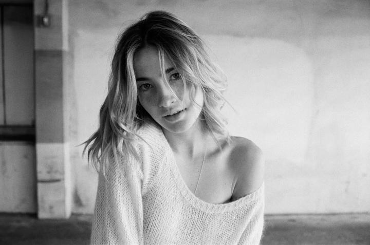 A woman with freckles and wavy hair looks at the viewer, wearing a loose-fitting sweater that exposes one shoulder.