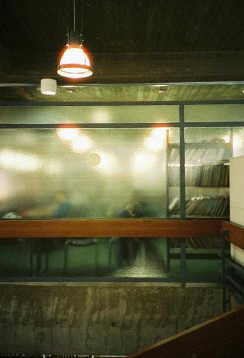 A hanging lamp illuminates a textured glass wall, behind which blurred figures and shelves of records are visible.