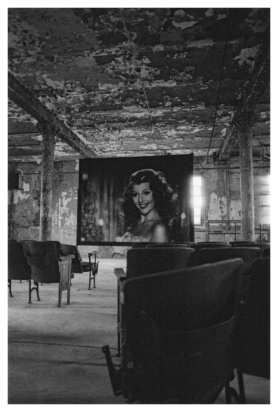 A black and white image shows a movie screen with a woman's face on it, surrounded by rows of empty seats in a dilapidated room.