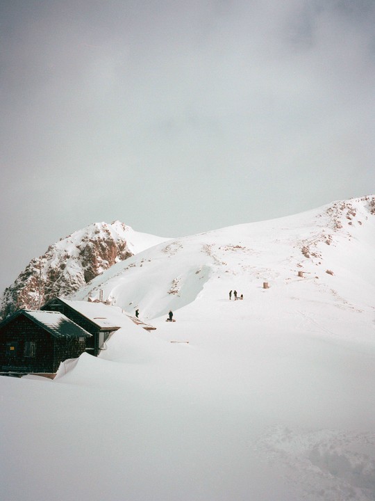 A snow-covered mountain landscape features several small buildings and a few people walking in the snow.