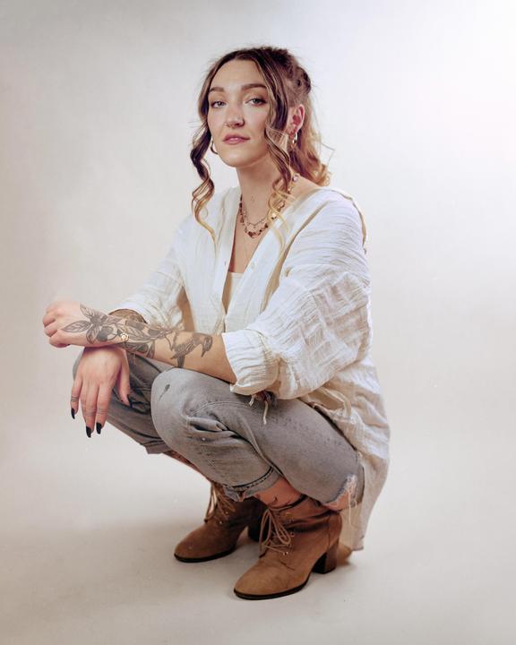 A woman with tattoos on her arms crouches while looking at the viewer.
