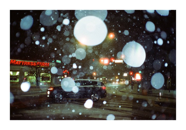 A black vehicle drives through a snowy street at night, with bright bokeh circles in the foreground and a Mattress Firm store in the background.