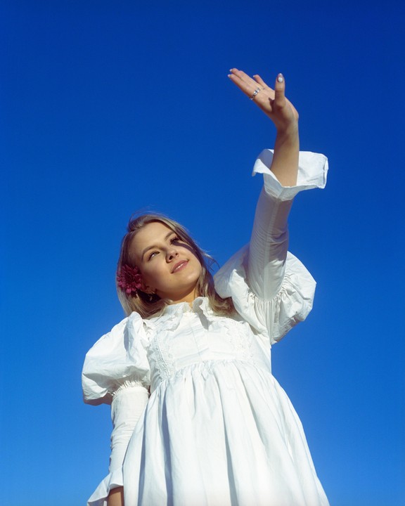 A woman in a white dress with a flower in her hair reaches her arm up towards a clear blue sky.