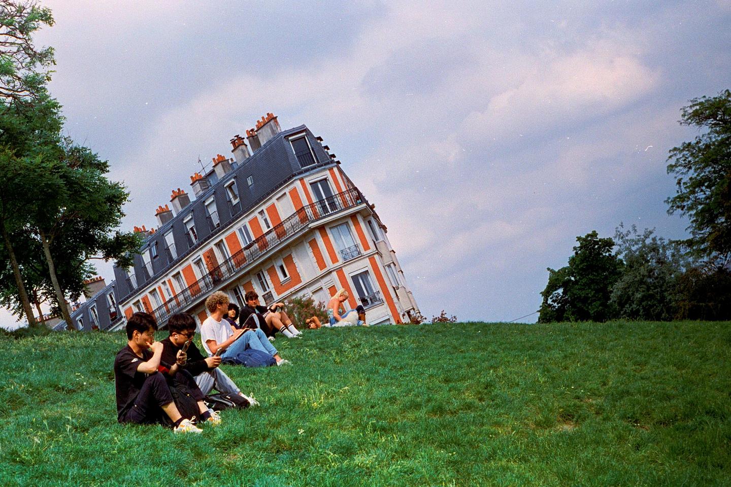 A group of people relax on a grassy hill with a building that appears to be sinking in the background.