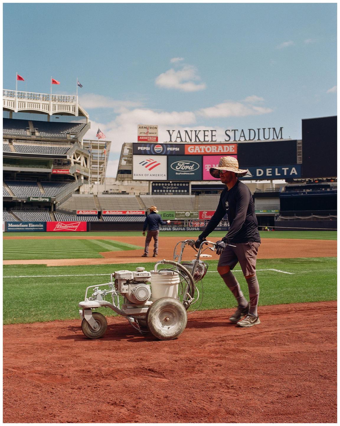 A man in a straw hat pushes a line marker on a baseball field with Yankee Stadium in the background.