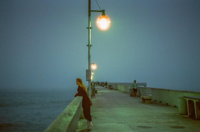 A woman leans on a railing on a pier at dusk, looking out at the hazy ocean.