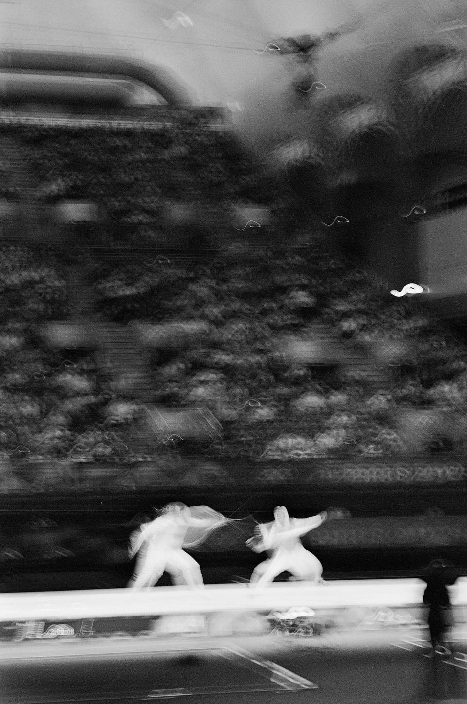 Two fencers are engaged in a match on a strip with spectators blurred in the background.