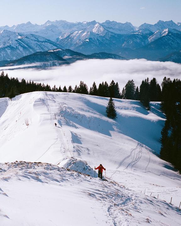 A person in a red jacket hikes up a snowy mountain with a valley of clouds and distant mountains in the background.