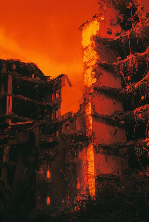 A partially demolished building stands against an orange sky, with rebar and debris visible.