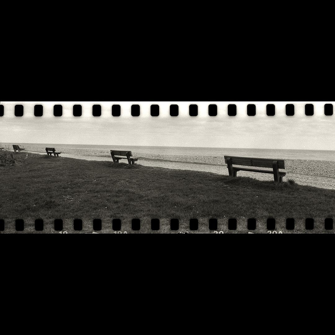 Three empty park benches overlook a calm sea on a grassy slope.