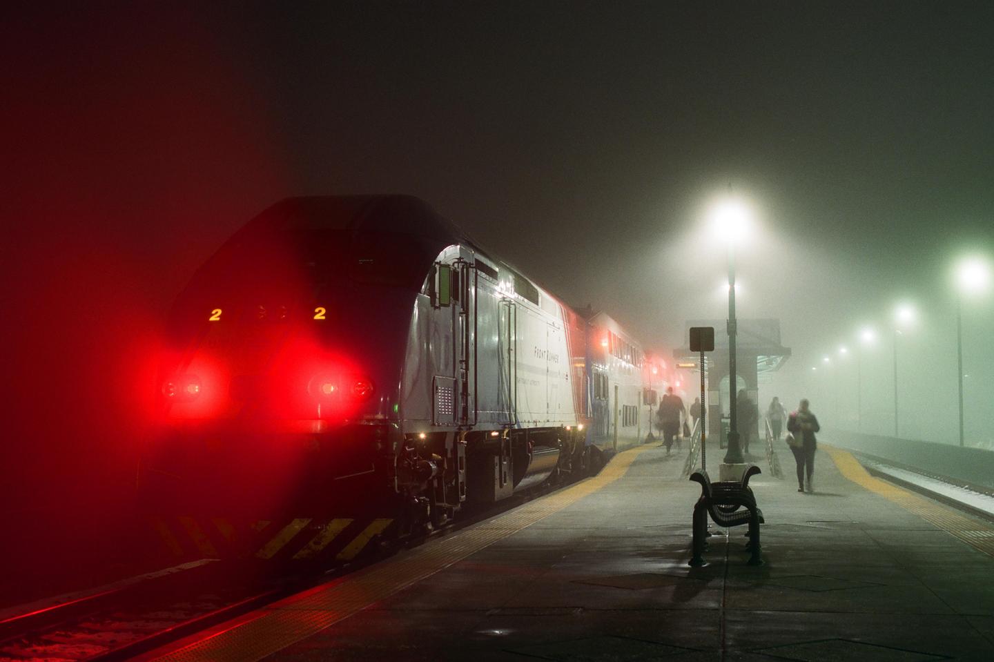 A train with bright red headlights is stopped at a station platform on a foggy night, with several people walking on the platform.