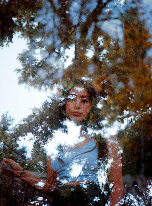 A woman's face and torso are visible through a double exposure of tree branches and sky.