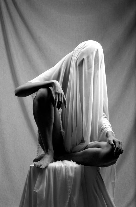 A person sits on a draped stool with a white sheet covering their head and body.